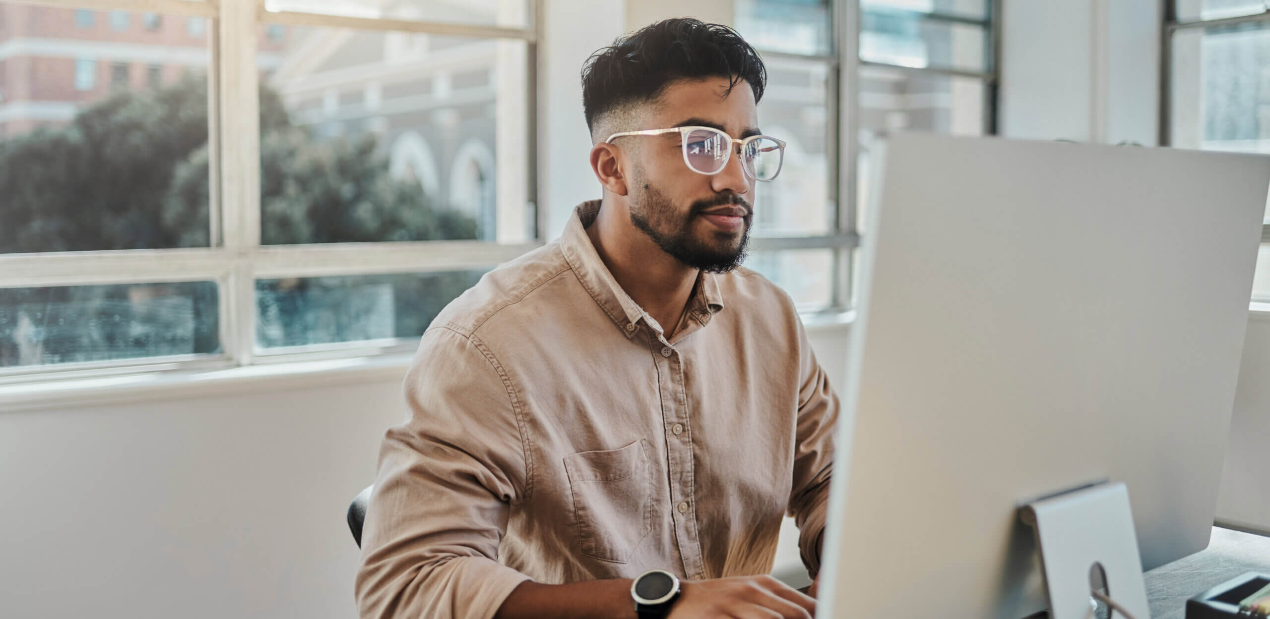 man with glasses, brown shirt looking at monitor at desk