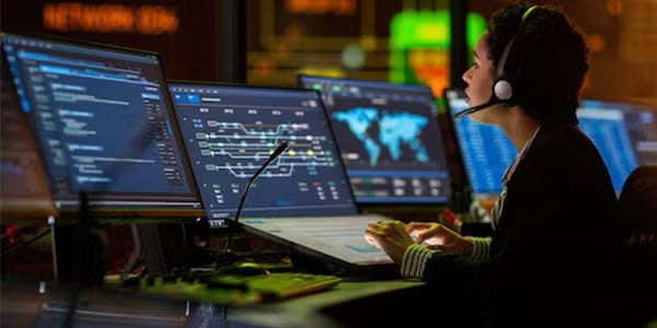 Multiethnic Female IT Technical Support Specialist Talking with a Client in Headphones and Working on Desktop Computer in Monitoring Control Room with Big Digital Screens with Server Blockchain Data.