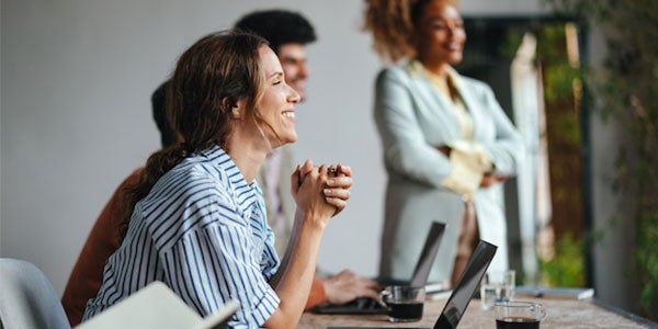 A side view of a smiling Caucasian entrepreneur using her laptop while attending a business meeting with a group of other employees.